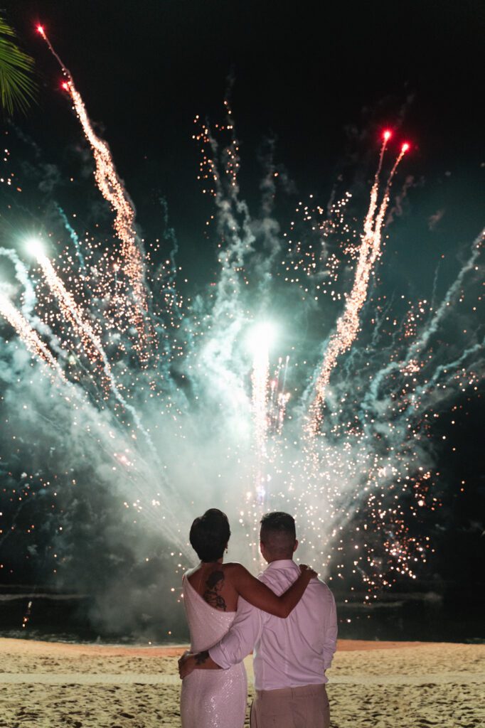 A couple shares a quiet moment beneath a sky lit by silent fireworks, creating a dramatic yet intimate ending to their wedding celebration
