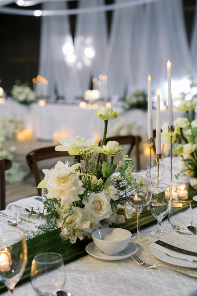 White wedding centerpiece with roses and greenery on a mirrored table runner, surrounded by tall candles and place settings