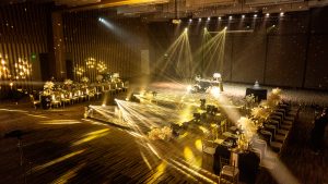 Grand ballroom reception setup at Vinpearl Nam Hội An with dramatic golden lighting, black tablecloths, gold chiavari chairs, feathered floral arrangements, and a central stage with moving light beams — Great Gatsby wedding theme by DNX Wedding.