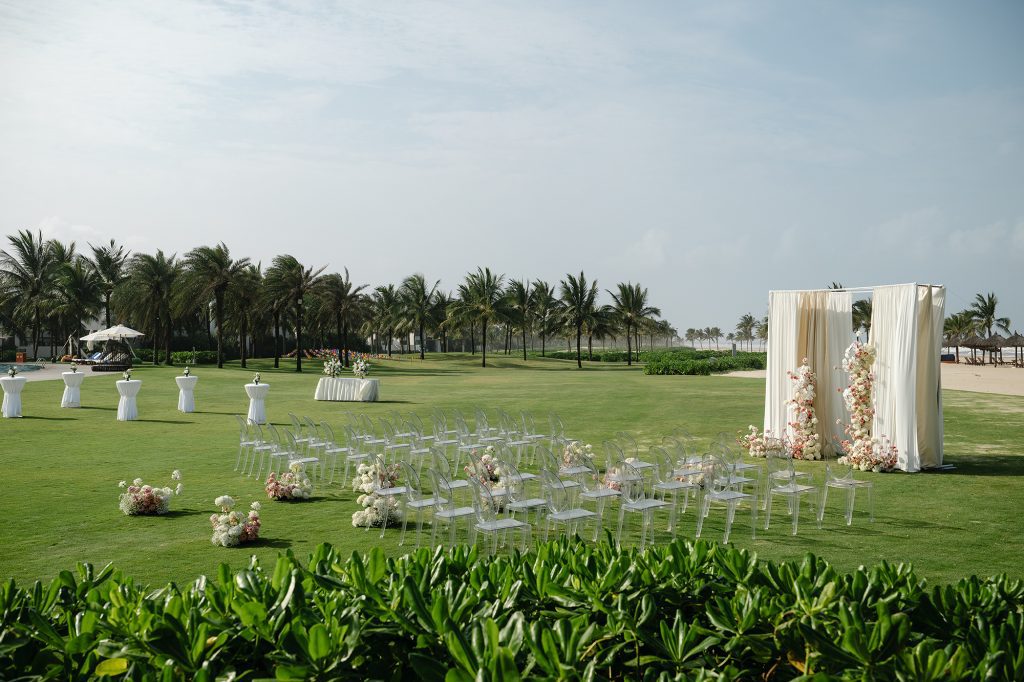 An elegant outdoor setup on a beachfront lawn for the wedding in Danang, with clear chairs, soft floral arrangements, and a ceremony backdrop surrounded by tropical greenery.