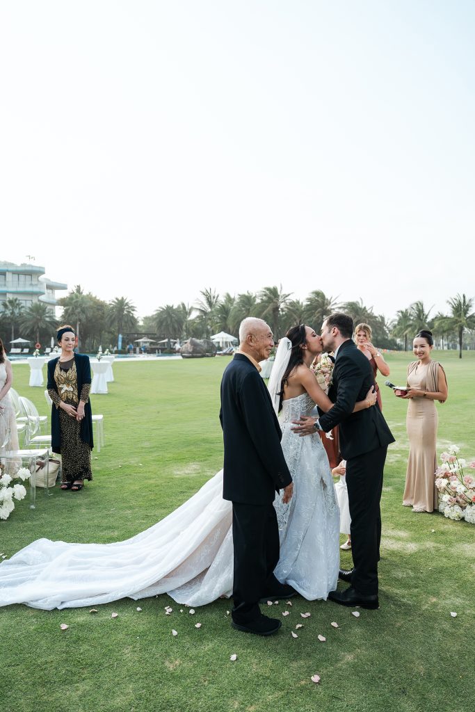 Bride and groom sharing their first kiss at the altar while the bride's father looks on at a beachfront wedding at Vinpearl Nam Hoi An