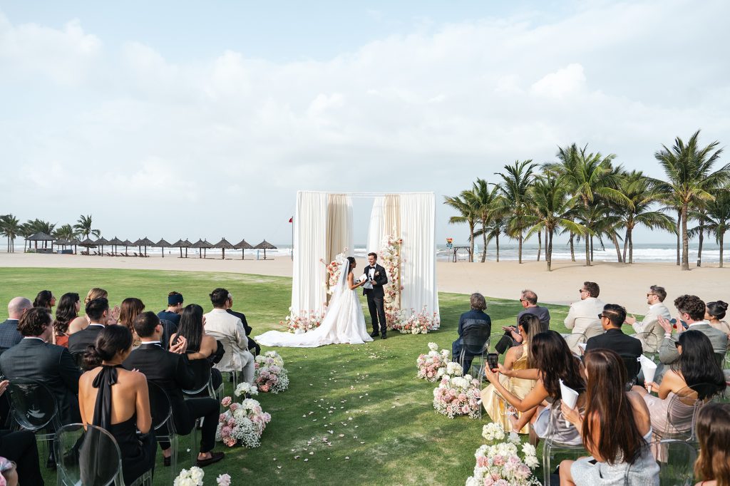 Couple exchanging vows at a beachfront wedding ceremony with ivory draped arch, blush floral aisle, and ocean backdrop at Vinpearl Nam Hoi An