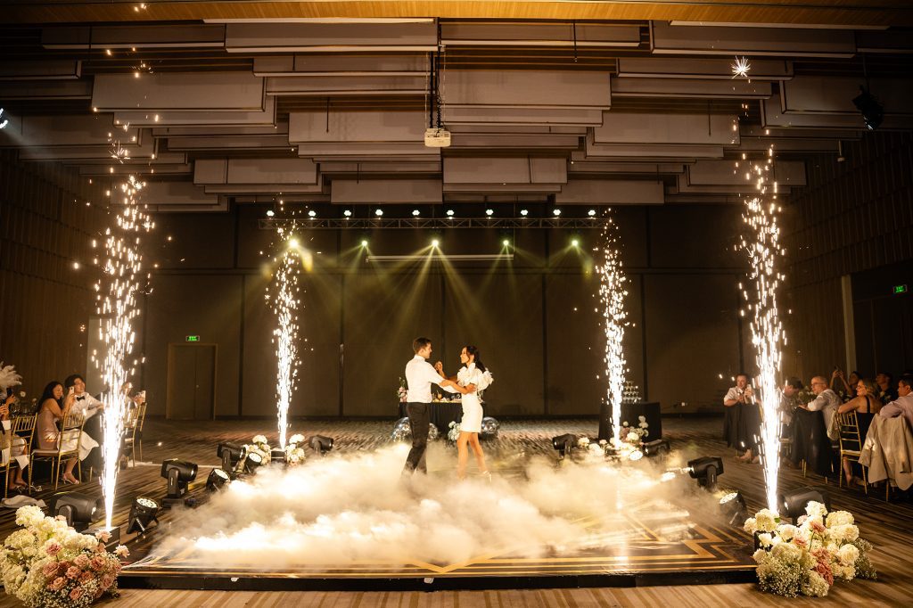 Bride and groom sharing their first dance on a fog-covered stage framed by four columns of cold sparkler fountains, guests watching from both sides, at the Great Gatsby-themed reception at Vinpearl Nam Hội An — planned by DNX Wedding.