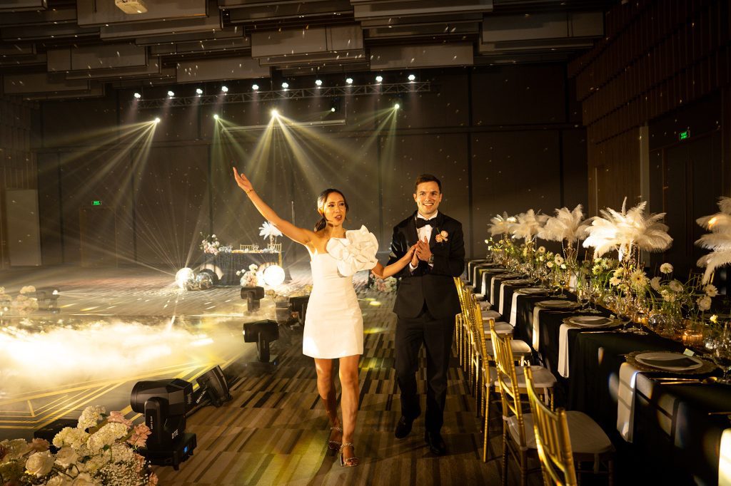 Bride in a white one-shoulder ruffle mini dress and groom in black tuxedo seeing the Great Gatsby-themed reception ballroom for the first time, with stage fog, golden spotlights, and feathered tablescape lining the aisle — Vinpearl Nam Hội An, planned by DNX Wedding.