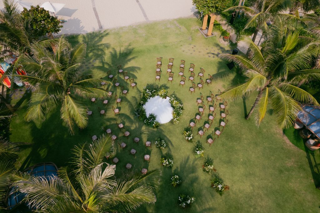 An aerial, high-angle view of an intimate, cozy wedding setup at Fusion Resorts & Villa Danang. Several wooden chairs are neatly arranged on a lush green lawn, with clear shadows cast by the surrounding tall palm trees in the warm daylight. A beach area with white umbrellas and the turquoise ocean are partially visible in the background.