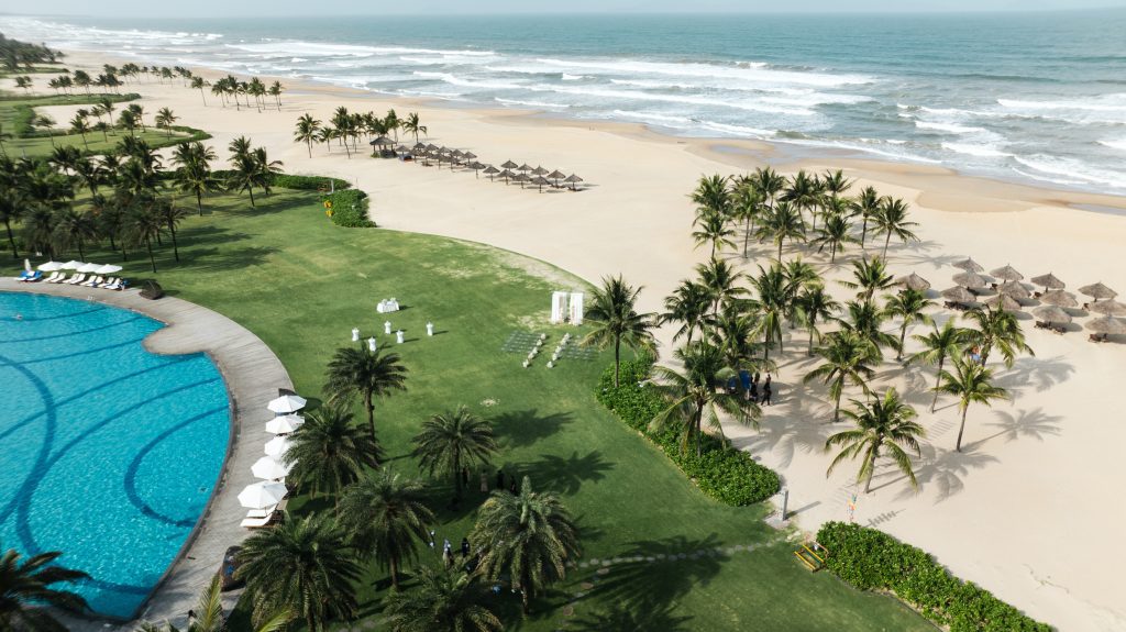 Aerial view of a beachfront resort with a large green lawn set for an outdoor wedding ceremony beside the pool and ocean