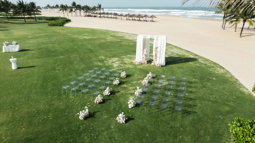 Aerial view of an outdoor beachside wedding ceremony setup with clear chairs, a floral arch, and palm trees along the Da Nang coastline