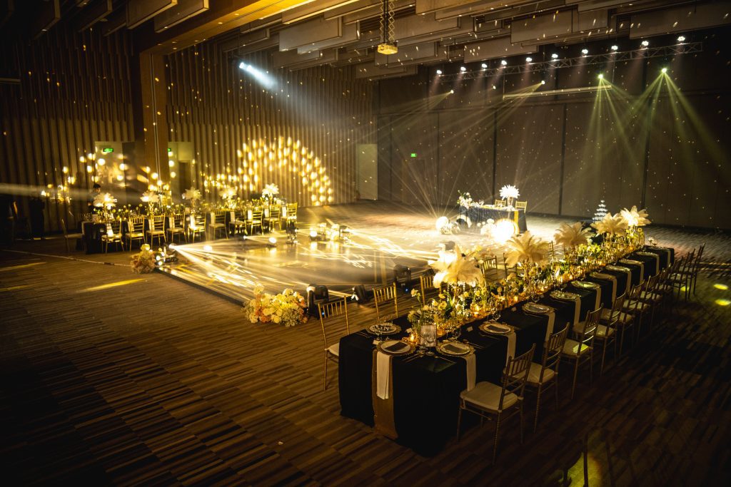 Elegant indoor wedding reception hall with dramatic lighting, long banquet table with black linens and white floral arrangements, and an illuminated dance floor