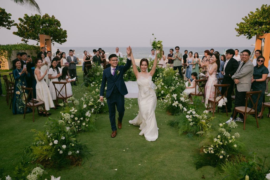 A bride and groom walk down a grass aisle lined with white flowers, holding hands and raising their arms in celebration. Guests on both sides cheer and clap under a clear sky, with the ocean softly visible in the background.