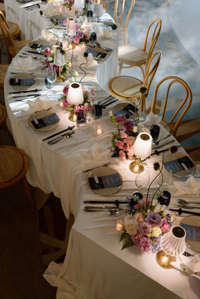 Elegant table setting with white linens, gold chairs, candles, and pastel floral centerpieces for a wedding reception