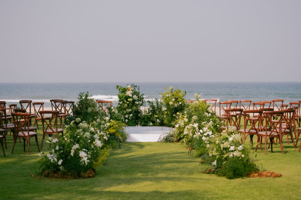 Outdoor wedding altar setup in Da Nang during peak sunny season, crescent-shaped floral arch with white flowers, wooden chairs facing the crystal-clear ocean