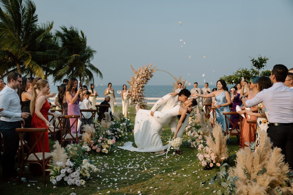 Bride and groom share a romantic dip kiss along a flower-lined aisle at an oceanside outdoor wedding, guests cheering and tossing petals, palm trees and pampas grass framing the scene.