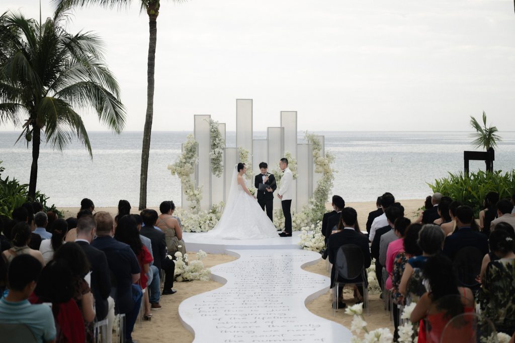 Bride and groom stand at a white floral altar during a beachfront wedding ceremony, with guests seated along a curved white aisle and the ocean in the background.