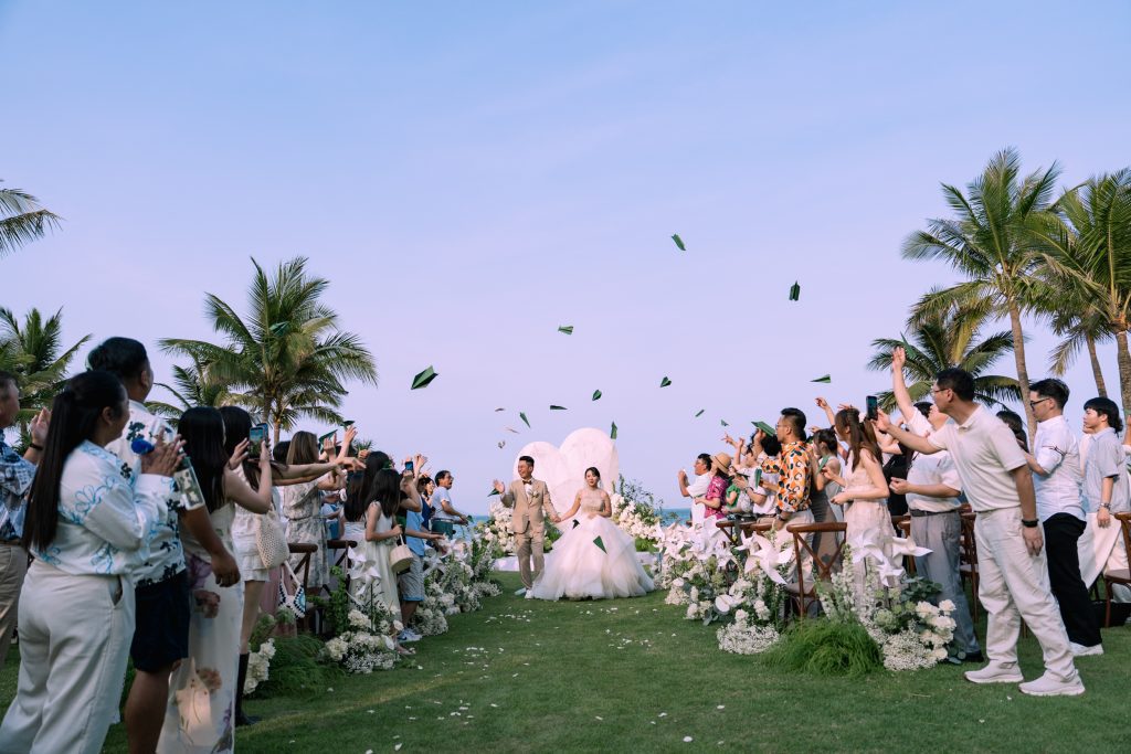 Sunny peak-season beach wedding in Danang - bride and groom walking down the grass aisle as guests toss paper pinwheels under clear blue skies