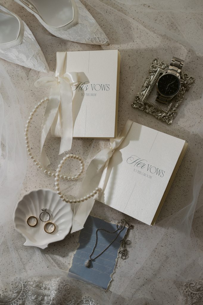 Wedding stationery flat lay with invitation cards, pearl bracelet, vintage frame, and blue gemstone pendant on a white sheer fabric background