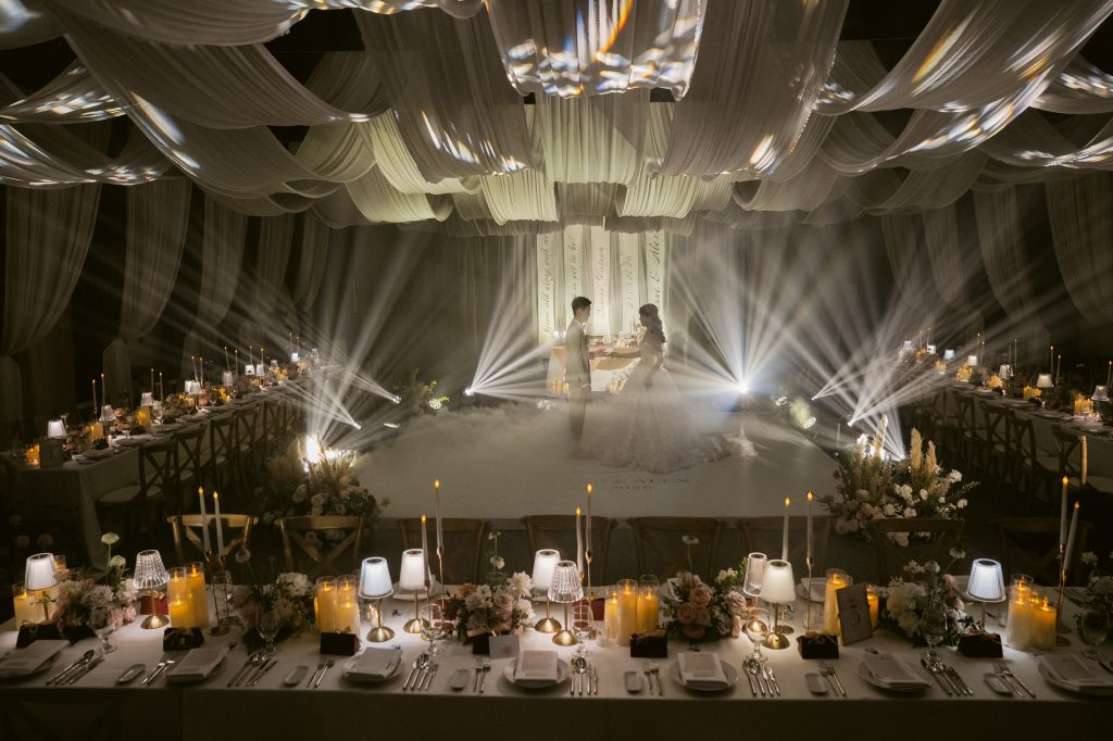 Evening wedding reception inside Naman Hall at Naman Retreat Danang, bride and groom sharing a first dance under dramatic spotlight beams, with long banquet tables lit by candles on either side.