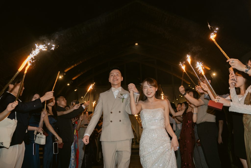 Bride and groom walking hand in hand through a sparkler send-off tunnel under Naman Hall's bamboo ceiling at Naman Retreat Danang, guests cheering on both sides.