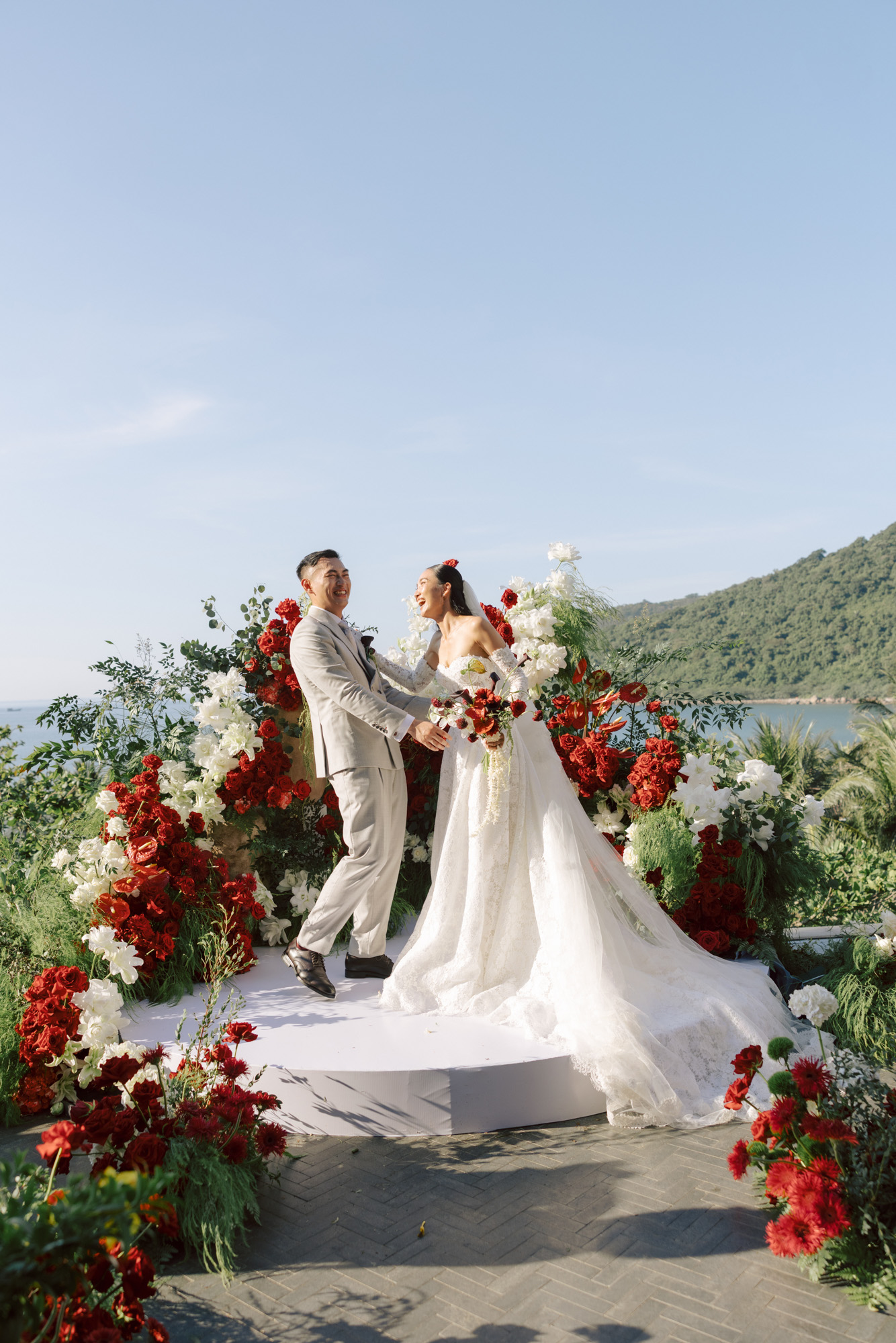 Alisa & Jeremiah's Cliff-View Wedding in Danang, bride and groom laughing together on a ceremony platform surrounded by red and white floral arches, with sea and mountain views in the background