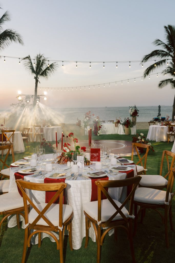 Da Nang beach wedding reception at dry-season sunset, table setting with red candles and fresh florals, string lights strung between palm trees, round dance floor overlooking the ocean