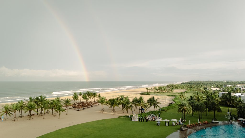 Aerial view of a Danang beach wedding during the rainy season, guests sheltering under umbrellas as a double rainbow arcs over the ocean and shoreline