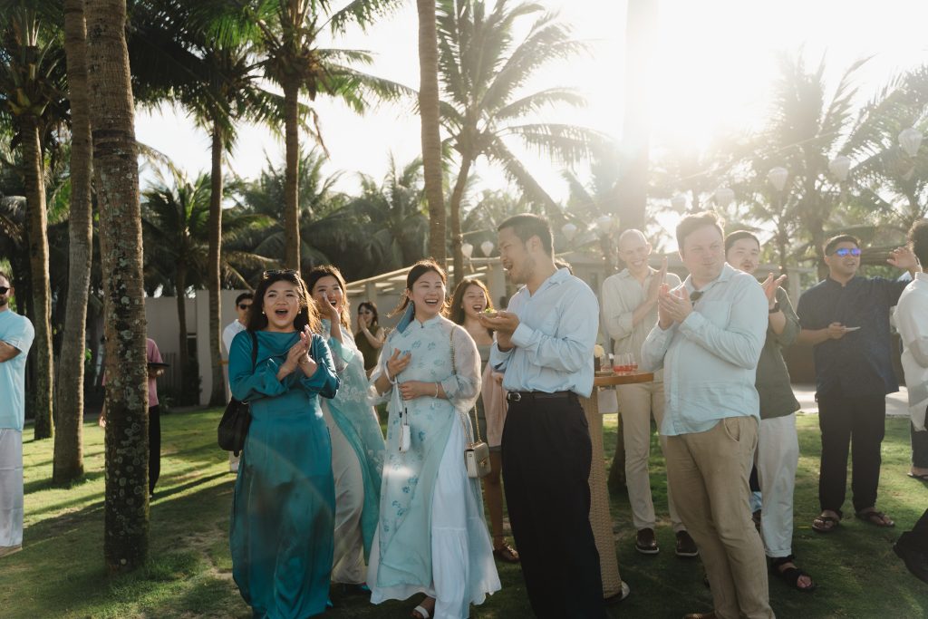 Wedding guests laughing and applauding outdoors under palm trees at golden hour during the dry season at Naman Retreat Danang, sunlight streaming through the coconut palms.