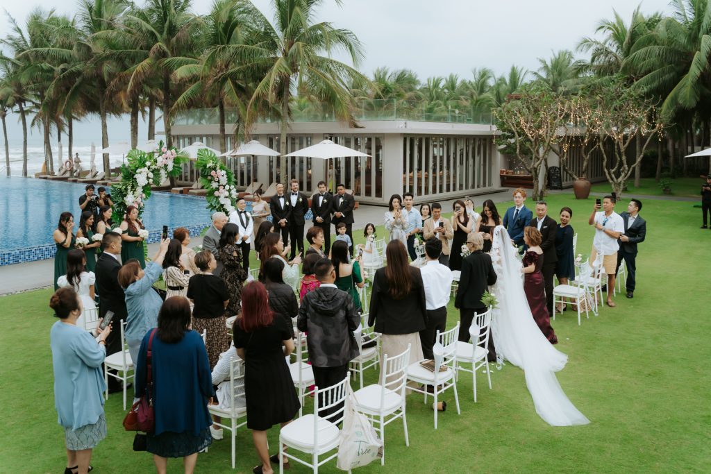 Outdoor wedding ceremony on Main Pool Lawn at Naman Retreat Danang, bride walking down a garden-lined aisle beside an infinity pool with palm trees and the resort's beachfront pavilion in the background.