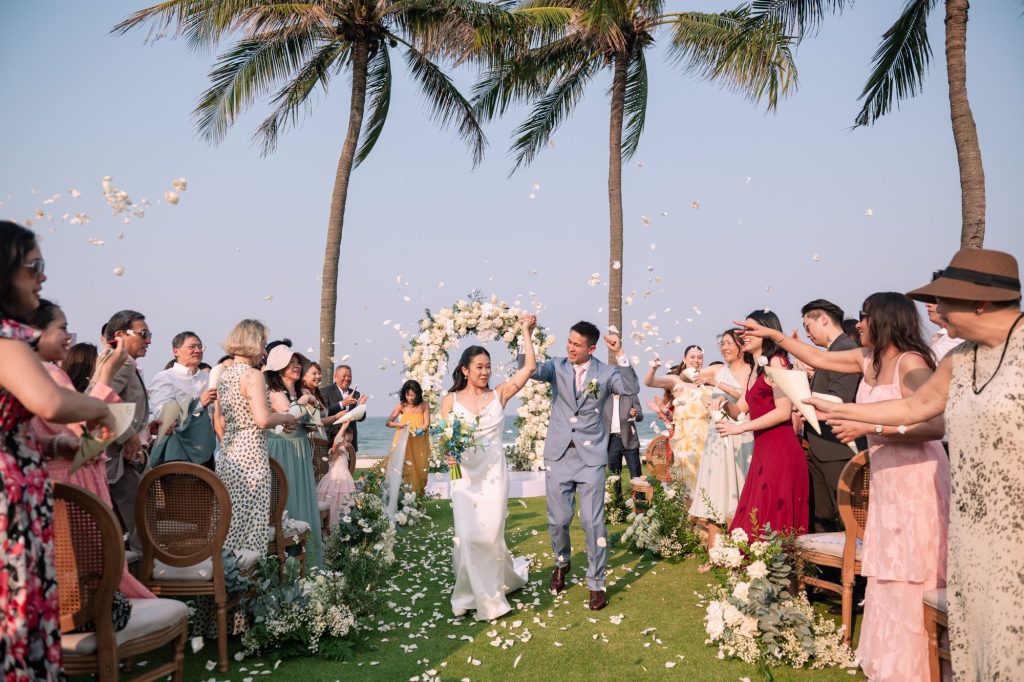 Bride and groom walking hand in hand down a petal-strewn aisle between palm trees under clear blue skies, guests cheering and tossing white petals on both sides, ocean visible in the background.